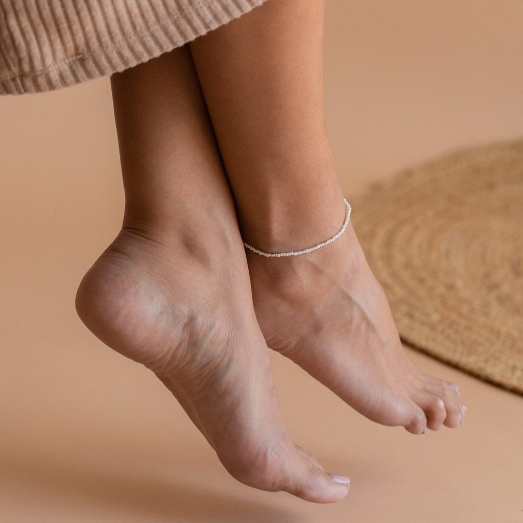 Ankle wearing a delicate, thin chain anklet dotted with tiny freshwater pearls, resting just above the curve of the heel, set against a warm beige background.
