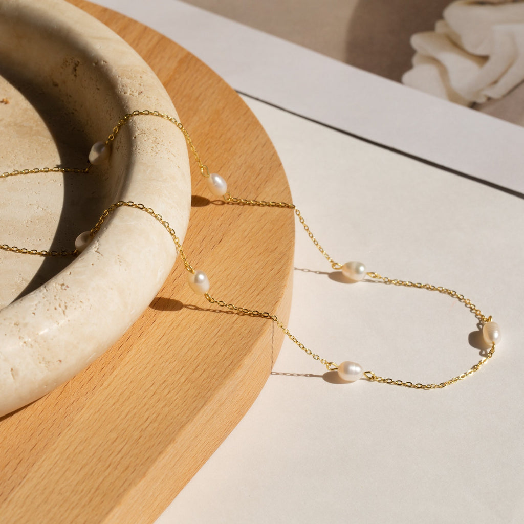 The Anna Pearl Station Necklace, featuring delicate gold with white pearls, is elegantly displayed on a round beige stone dish atop a wooden surface—perfect as a Mother’s Day or bridesmaids gift.