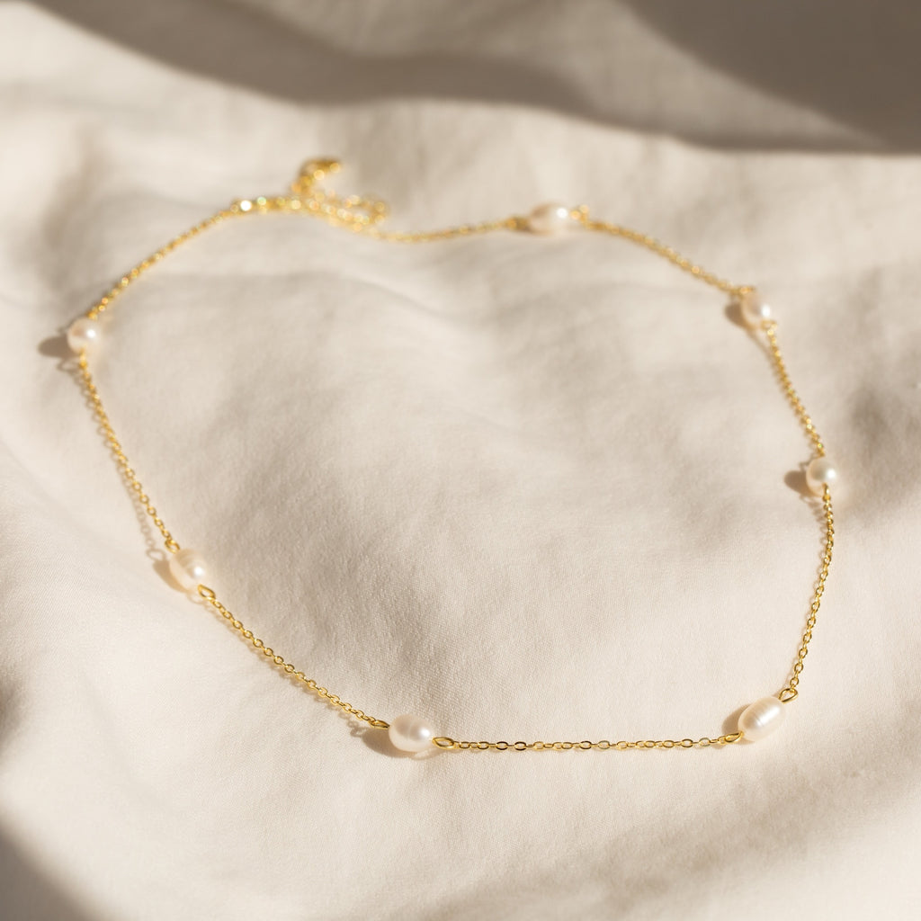 Gold chain necklace with evenly spaced oval white pearls, displayed on a soft cream fabric background.
