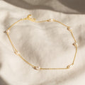 Gold chain necklace with evenly spaced oval white pearls, displayed on a soft cream fabric background.
