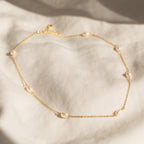 Gold chain necklace with evenly spaced oval white pearls, displayed on a soft cream fabric background.

