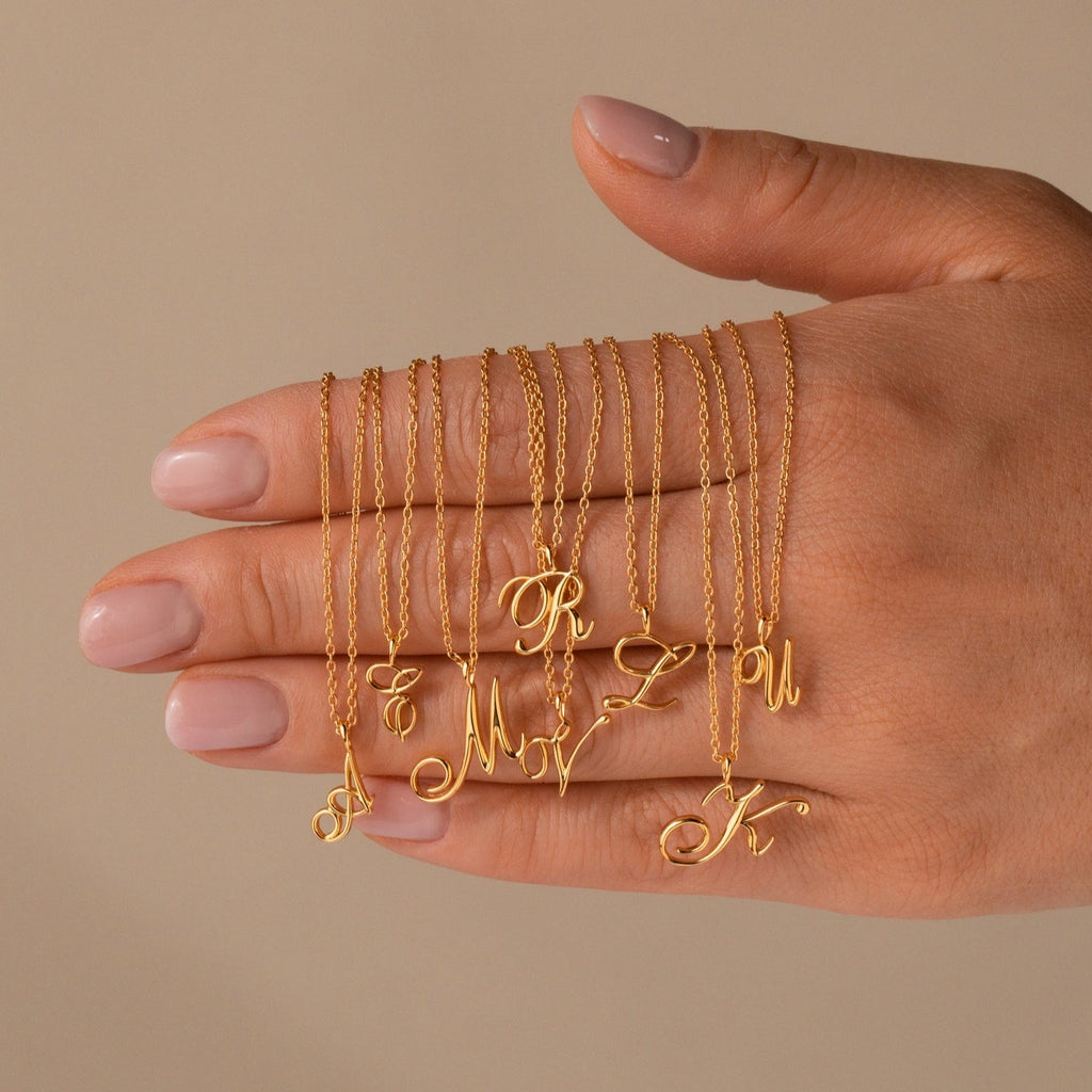 A collection of gold necklaces with cursive initial pendants draped over a woman's hand. The delicate chains feature script-style letters including A, T, M, R, V, Z, U, and K, showcasing a graceful and personalized jewelry style against a neutral background.