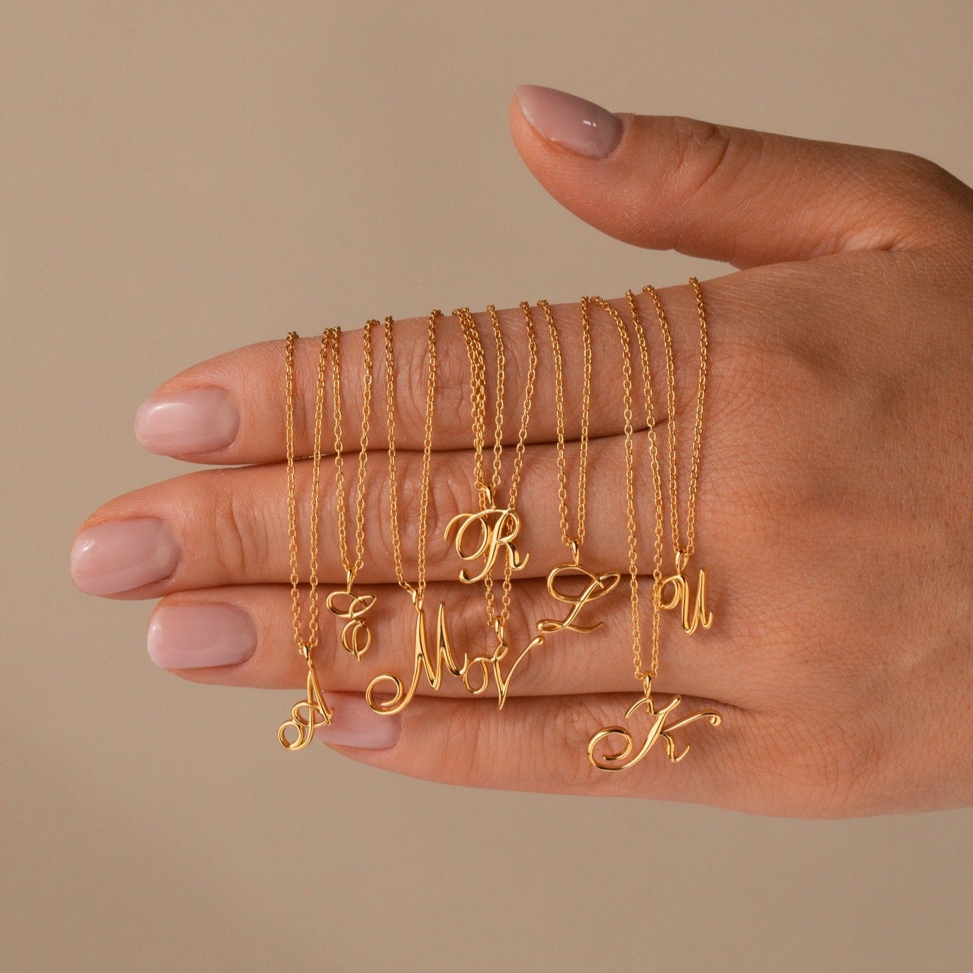 A collection of gold necklaces with cursive initial pendants draped over a woman's hand. The delicate chains feature script-style letters including A, T, M, R, V, Z, U, and K, showcasing a graceful and personalized jewelry style against a neutral background.