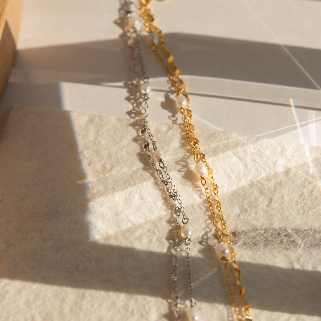Close-up of two Magnolia Pearl Wrap Bracelets—one in gold and one in silver—laid side by side on a textured cream surface, with soft natural sunlight highlighting the contrast between the warm gold and cool silver chains accented by small freshwater pearls.