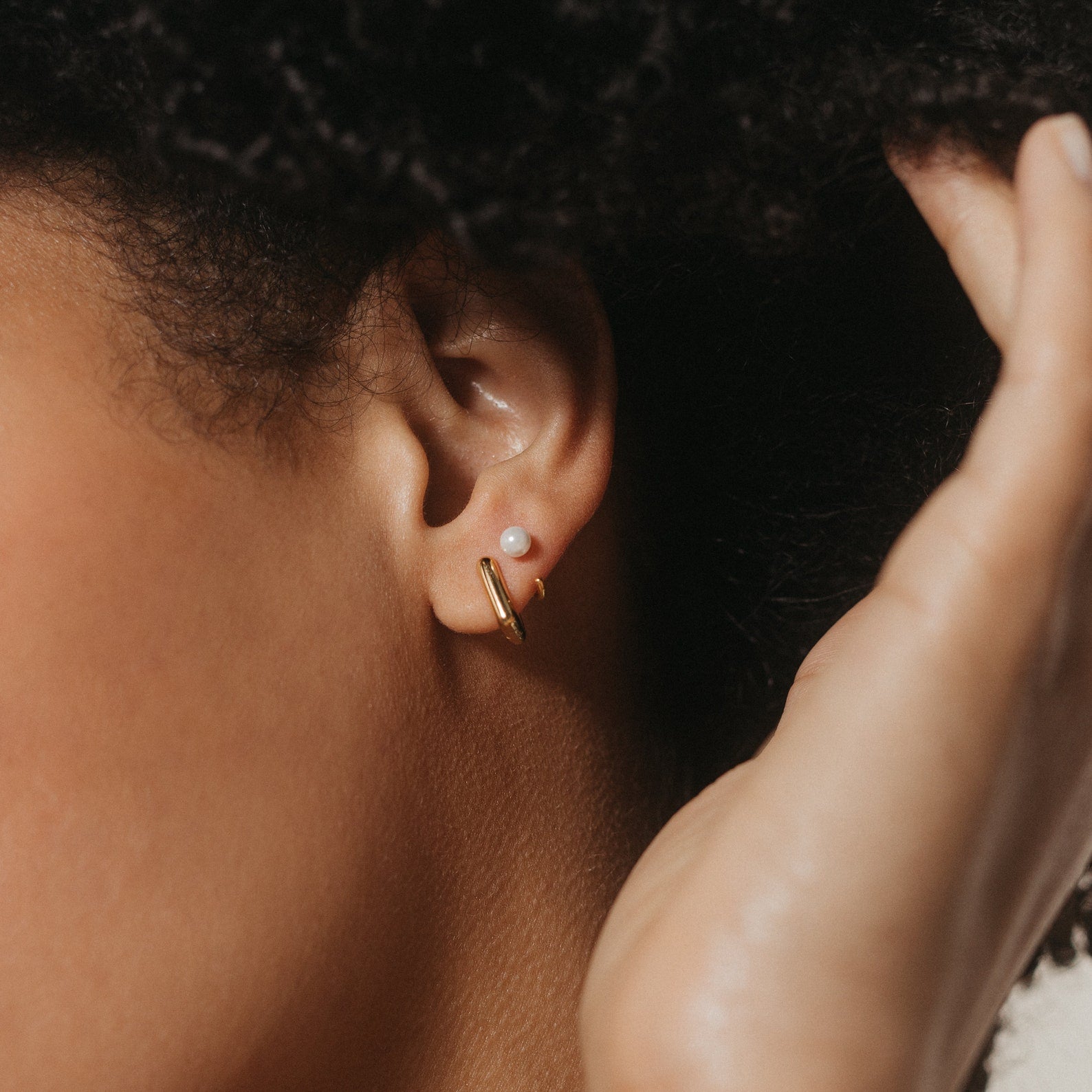 A close-up of a person's ear wearing a pearl stud and a minimal accessory: the contemporary Kelsey Link Earrings in gold.