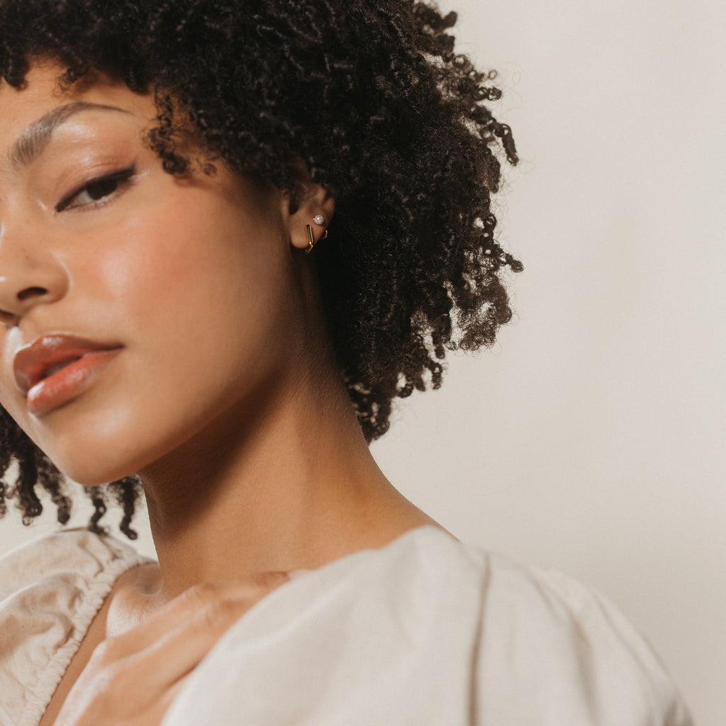 Woman with curly hair wearing a cream top looks at the camera, hand near collarbone, showcasing the Kelsey Link Earrings—a minimal rectangle geometric huggie—against a neutral background.