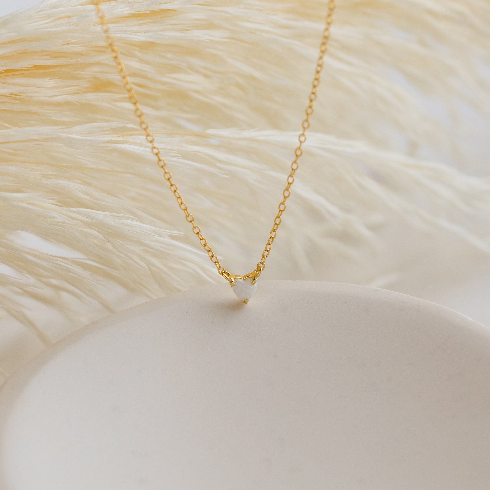 The Madeline Opal Heart Necklace features a delicate gold chain with a small heart-shaped opal pendant, elegantly displayed on a white surface with a soft, feathery background.