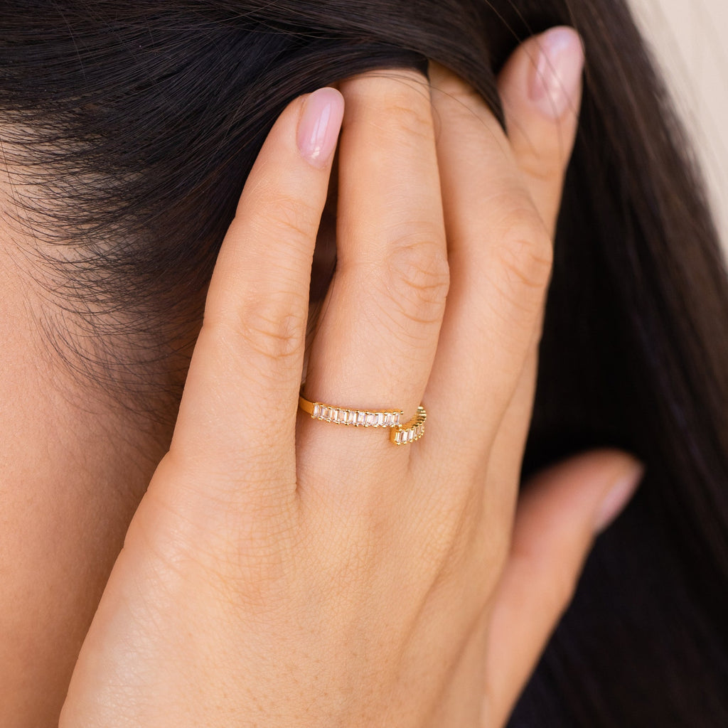 A hand with light pink nails wears the delicate Elora Baguette Wrap Ring, featuring sparkling diamond crystal baguettes, while gently touching dark brown hair.