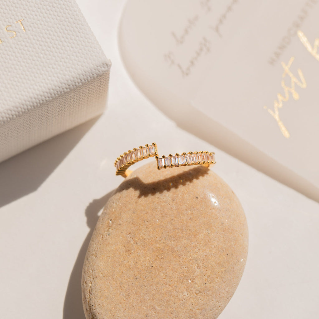 The Elora Baguette Wrap Ring, adorned with diamond crystal baguettes, rests on a smooth beige stone beside a box and a card.