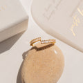 The Elora Baguette Wrap Ring, adorned with diamond crystal baguettes, rests on a smooth beige stone beside a box and a card.