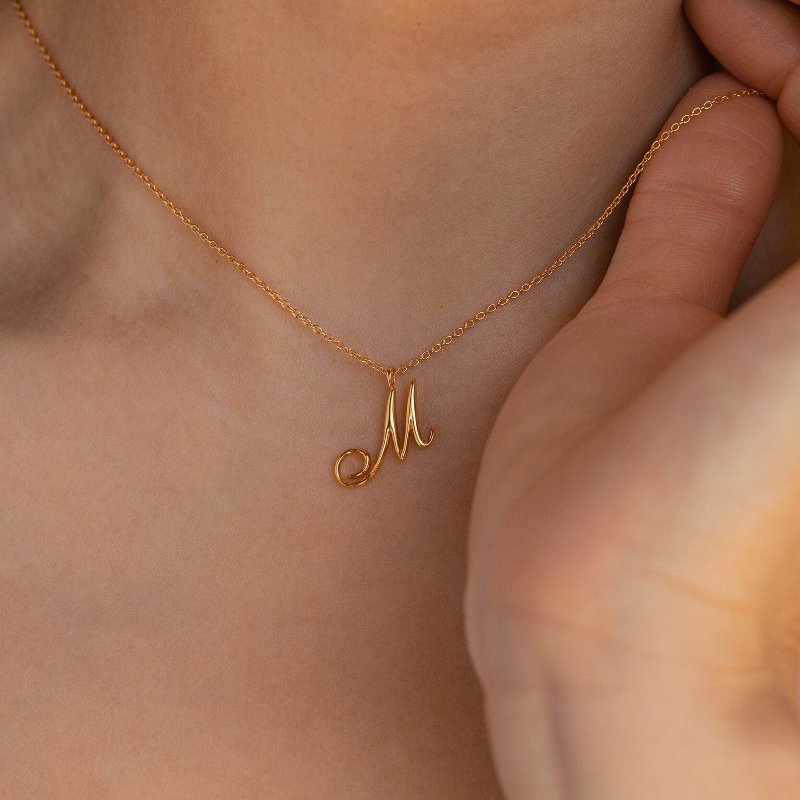The Davina Letter Necklace, featuring a gold M pendant, sits elegantly on a person’s neck with their hand near the chain—a perfect romantic gift.