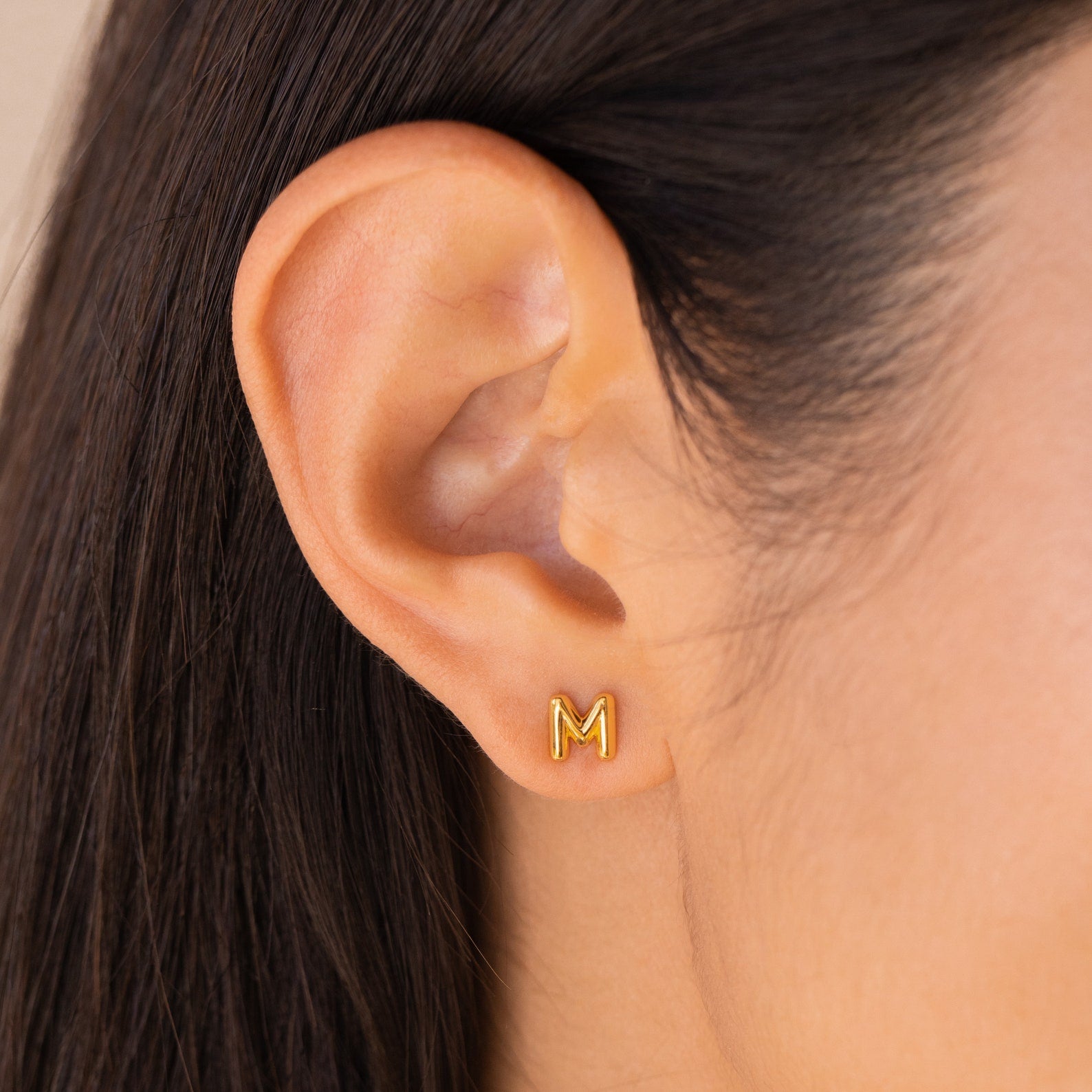 Close-up of an ear wearing Bubble Letter Studs, gold stud earrings shaped like the letter M—ideal as initial charm earrings or a unique bridesmaid gift.