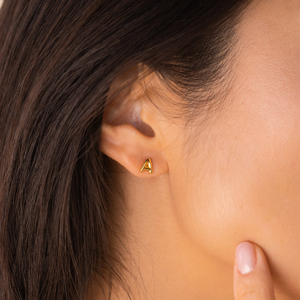 Close-up of a woman's ear adorned with a gold Bubble Letter Stud, perfect for personalizing your look or gifting to bridesmaids. Her finger rests on her cheek, drawing attention to the delicate earring.