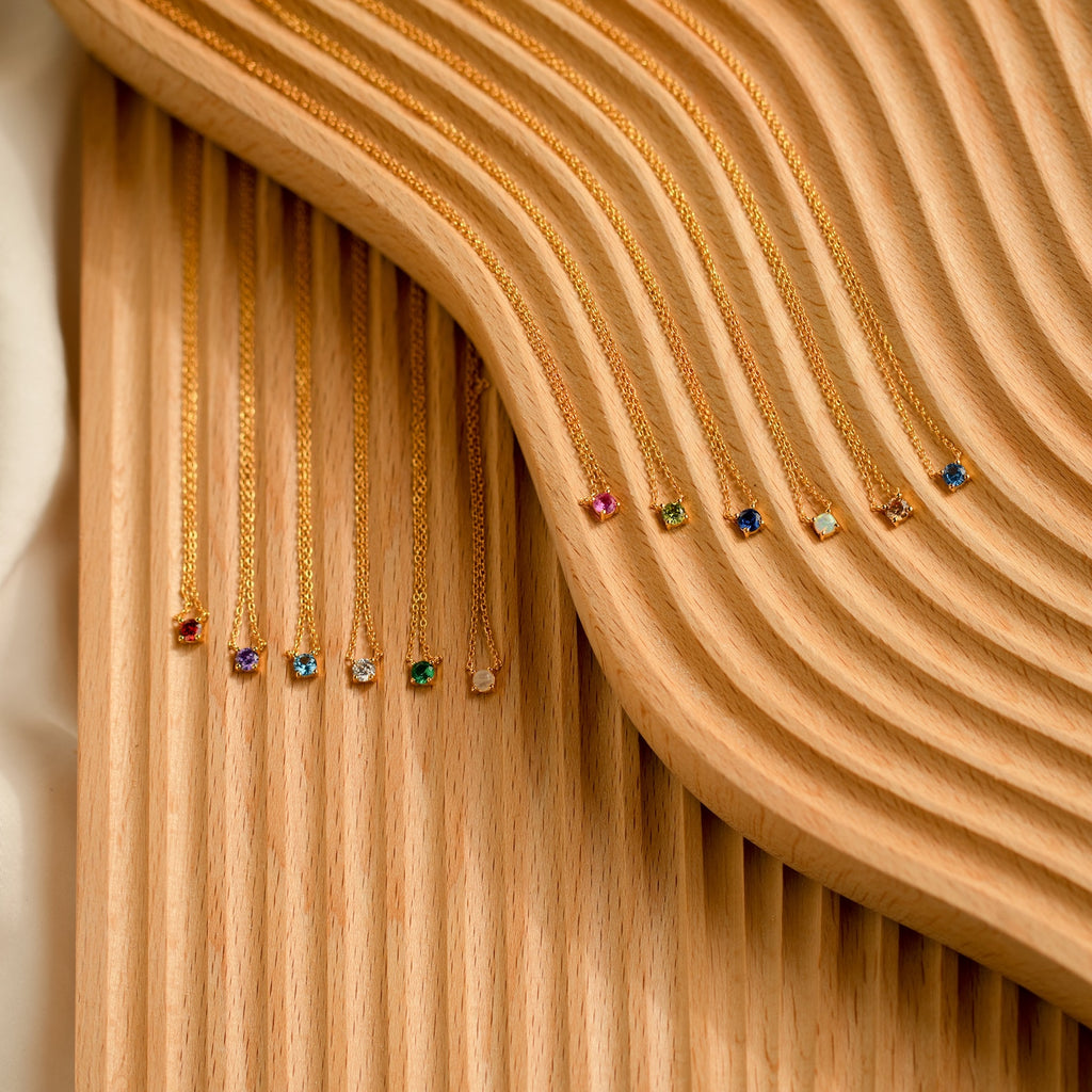 The Dainty Birthstone Necklace, featuring colorful gemstone pendants for each birth month, is displayed on a wavy wooden surface.
