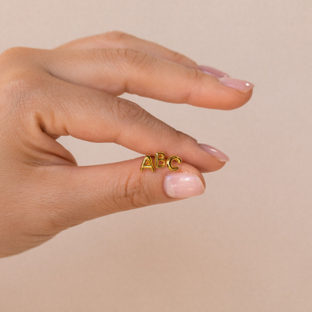 A hand holds small gold "ABC" Bubble Letter Studs—ideal inspiration for personalized or initial earrings, making a thoughtful bridesmaid gift against a neutral background.