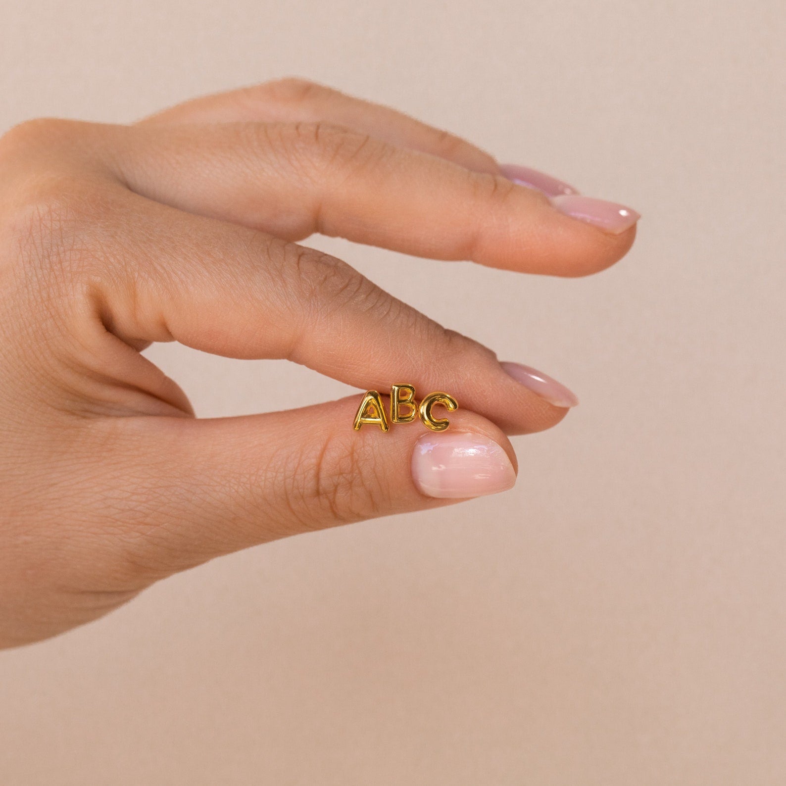A hand holds small gold "ABC" Bubble Letter Studs—ideal inspiration for personalized or initial earrings, making a thoughtful bridesmaid gift against a neutral background.