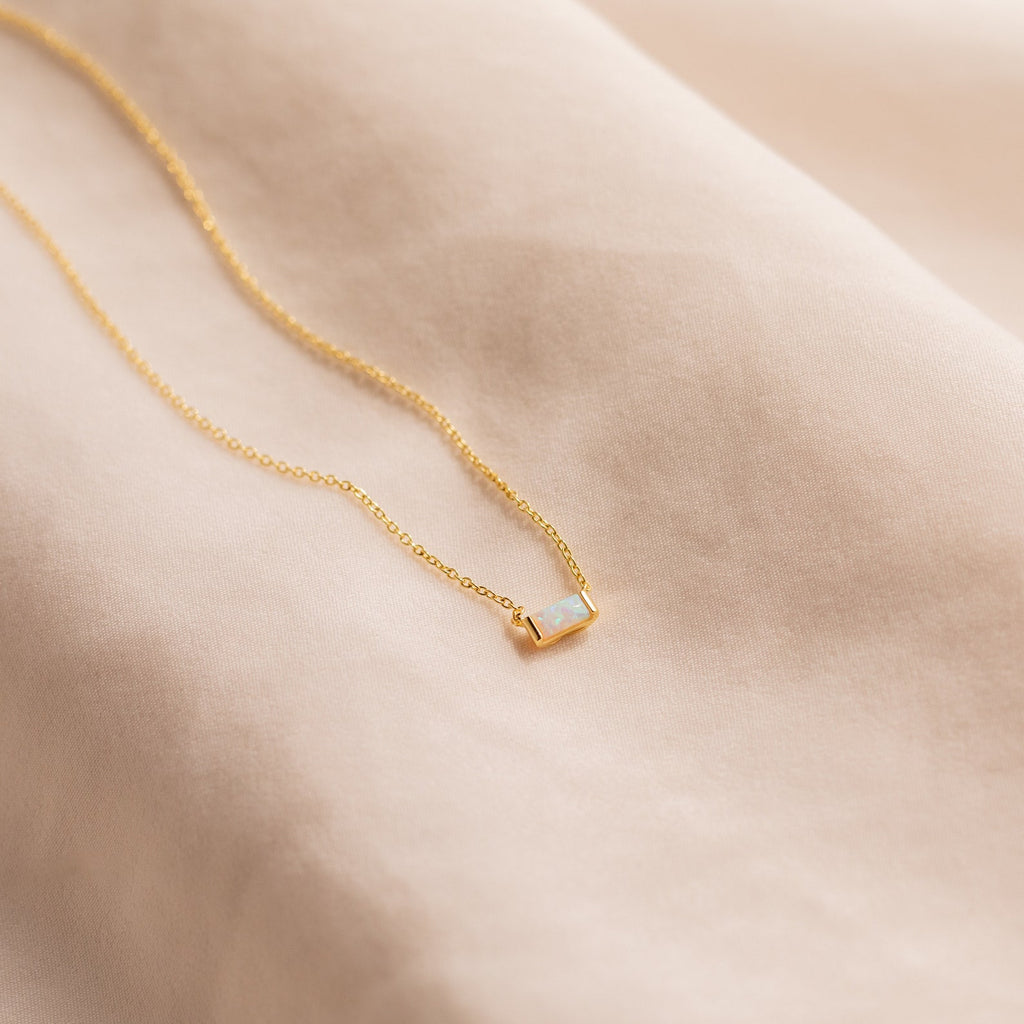 A gold Emery Birthstone Necklace featuring a soft opal rectangular gemstone, laid on beige fabric.