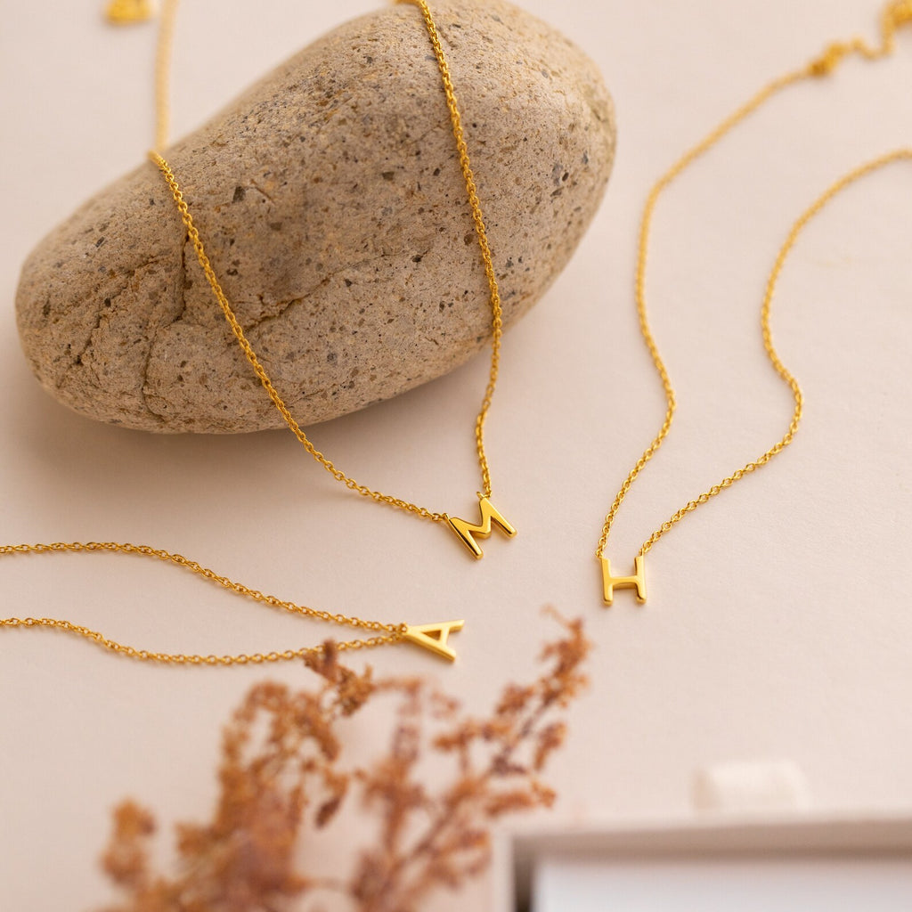 Three gold Dainty Initial Necklaces, each featuring a letter pendant (A, H, M), rest on a beige surface with a smooth stone—a customizable Dainty Initial Necklace perfect for a thoughtful Christmas gift.