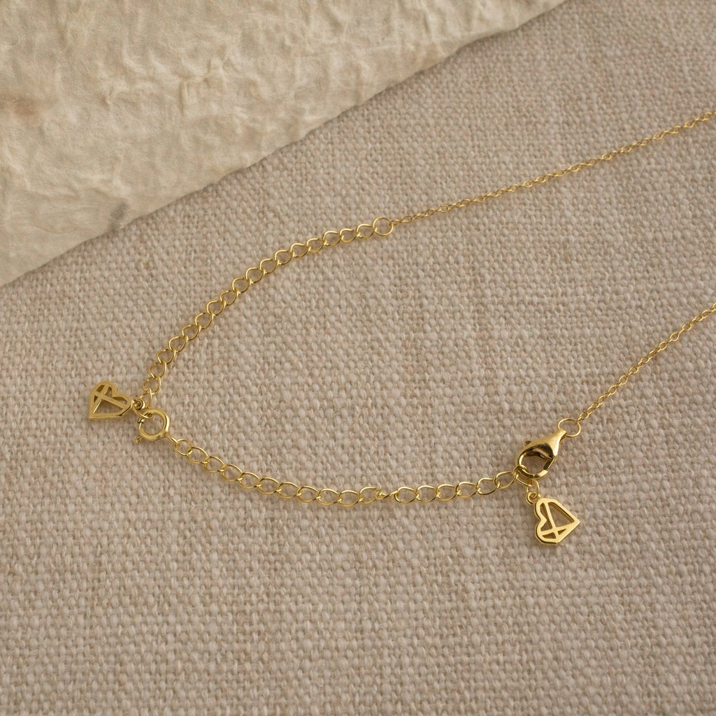 The Chain Extender is shown on a beige fabric surface, attached to a gold chain necklace with small geometric triangle charms, and offers an adjustable fit.