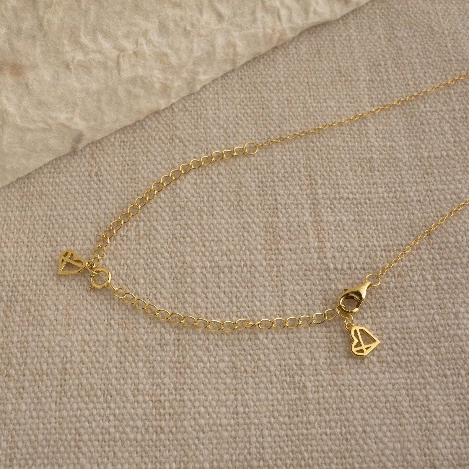 The Chain Extender is shown on a beige fabric surface, attached to a gold chain necklace with small geometric triangle charms, and offers an adjustable fit.