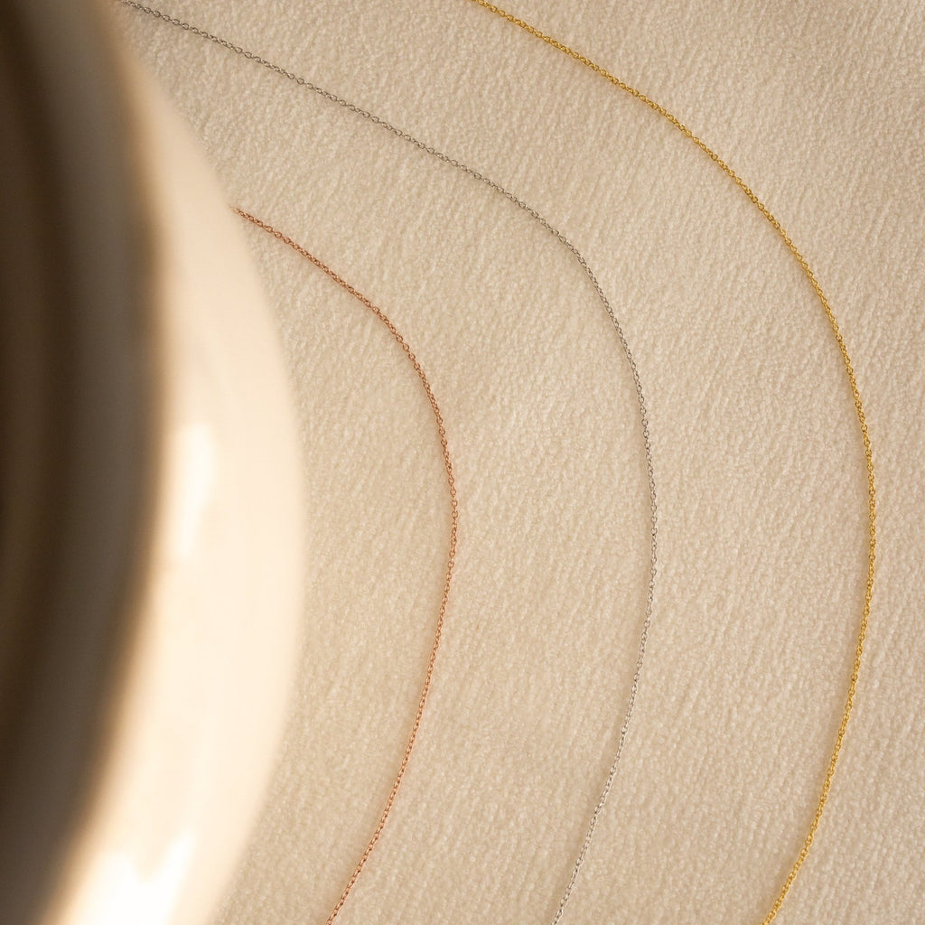 The Dainty Chain Necklace, shown in gold, silver, and rose gold, curves across a beige textured surface with a blurred object to the left for a refined minimal look.