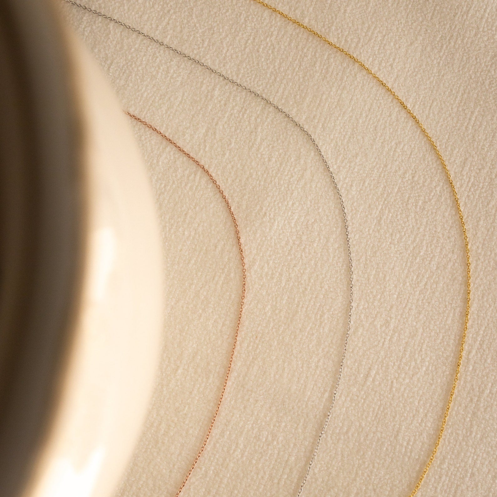 The Dainty Chain Necklace, shown in gold, silver, and rose gold, curves across a beige textured surface with a blurred object to the left for a refined minimal look.