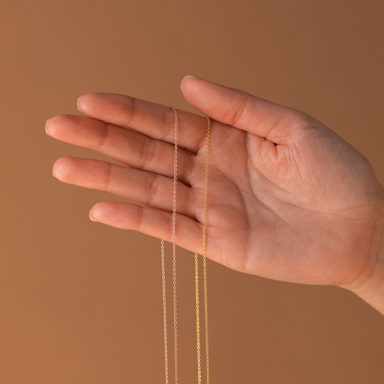 A hand holds two Dainty Chain Necklaces in silver and gold, ideal for a minimal look or stylish necklace stack, set against a neutral brown background.