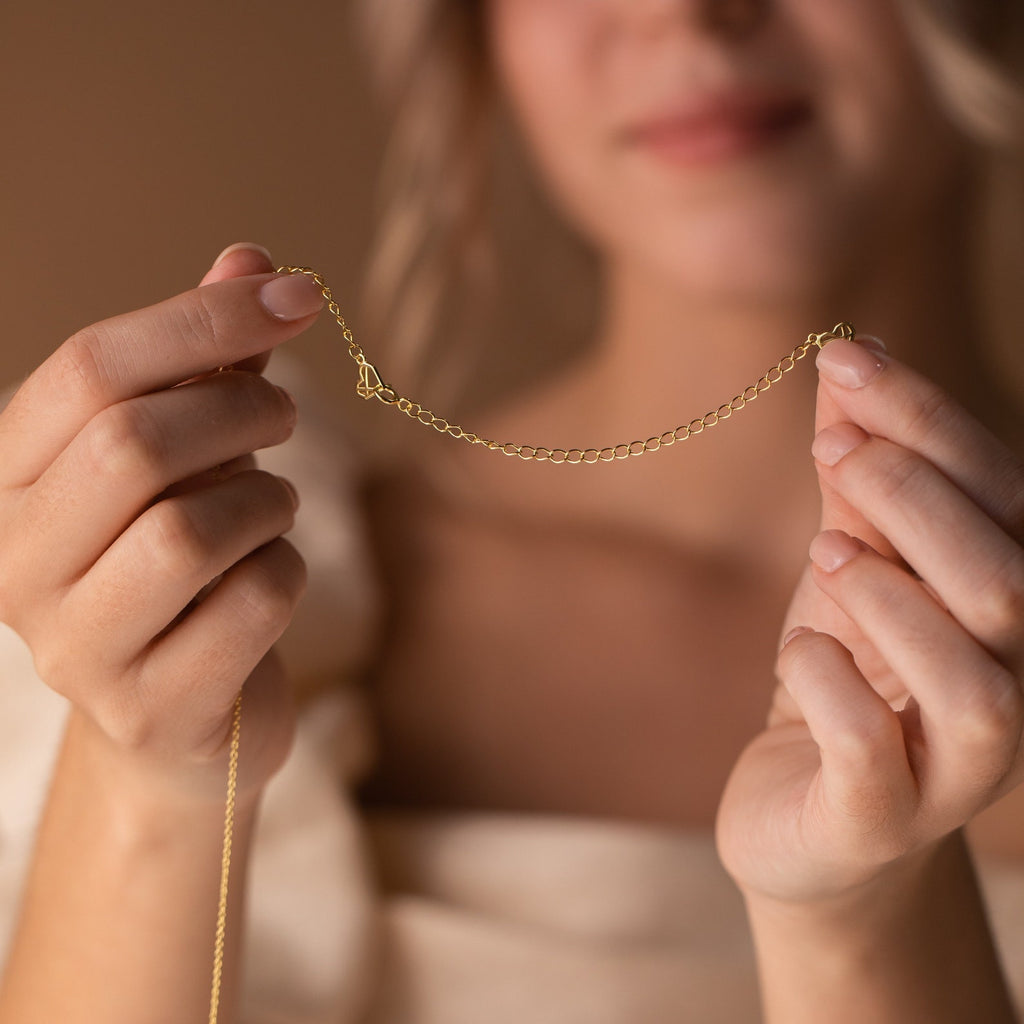 A woman holds a delicate gold Chain Extender necklace with both hands, highlighting its subtle added length; her face remains softly out of focus.