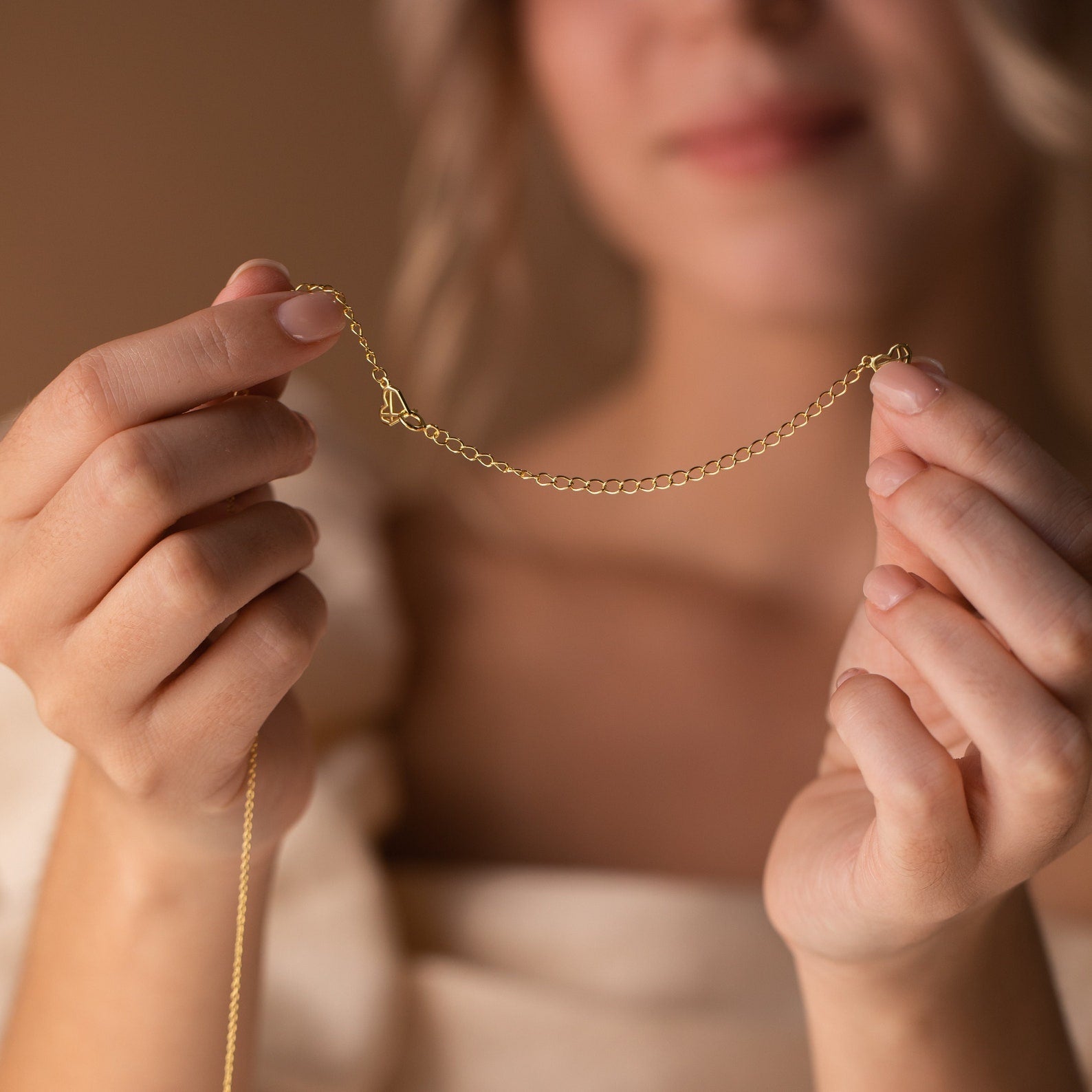 A woman holds a delicate gold Chain Extender necklace with both hands, highlighting its subtle added length; her face remains softly out of focus.