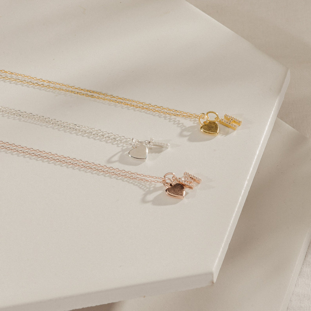 The Pave Initial Heart Necklace, featuring delicate heart and tag charms in gold, silver, and rose gold tones, is beautifully displayed on a white background.