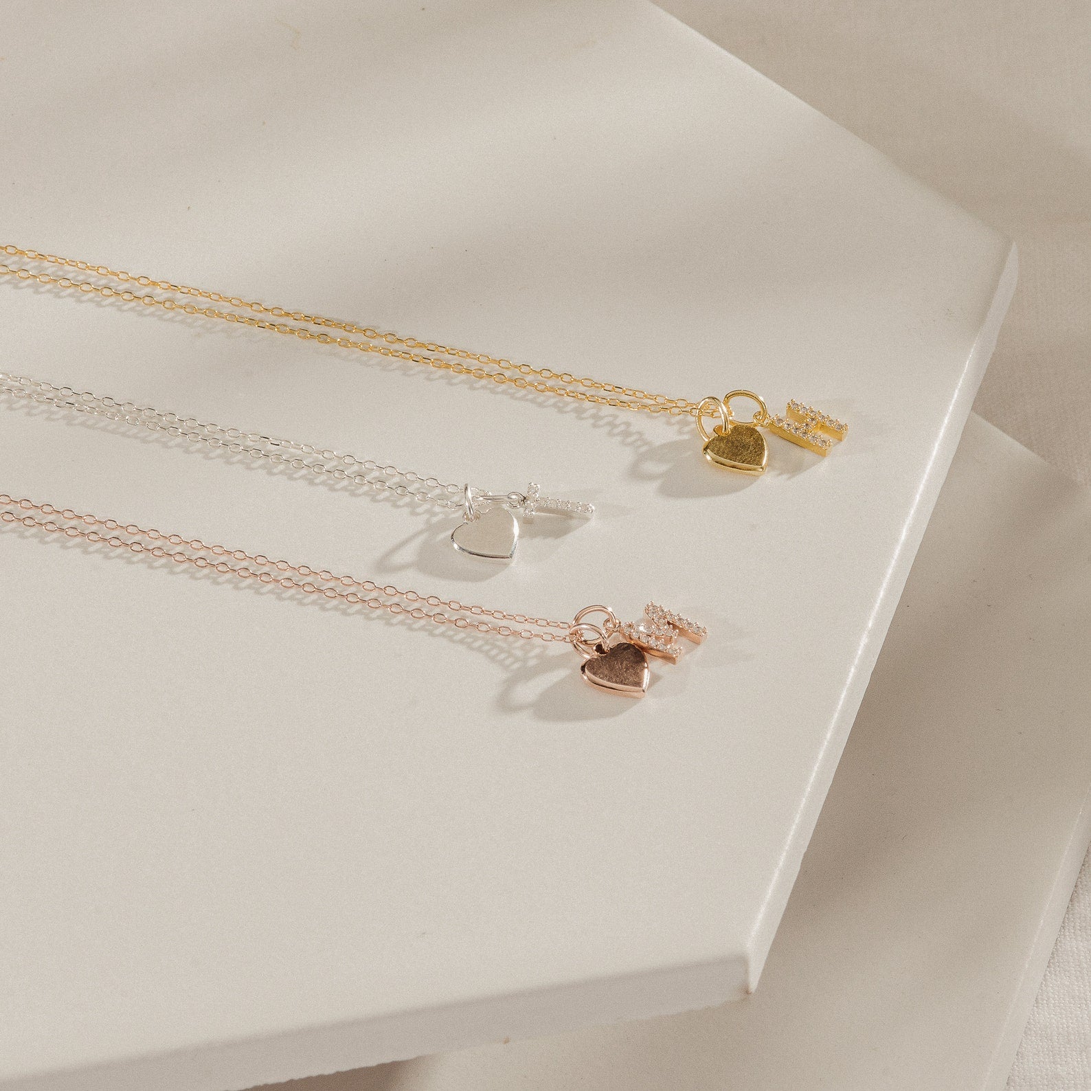 The Pave Initial Heart Necklace, featuring delicate heart and tag charms in gold, silver, and rose gold tones, is beautifully displayed on a white background.