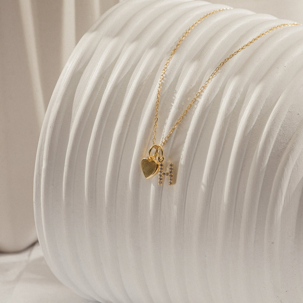 Pave Initial Heart Necklace featuring a gold heart charm and initial pendant with rectangular accents, elegantly displayed on a textured white surface.