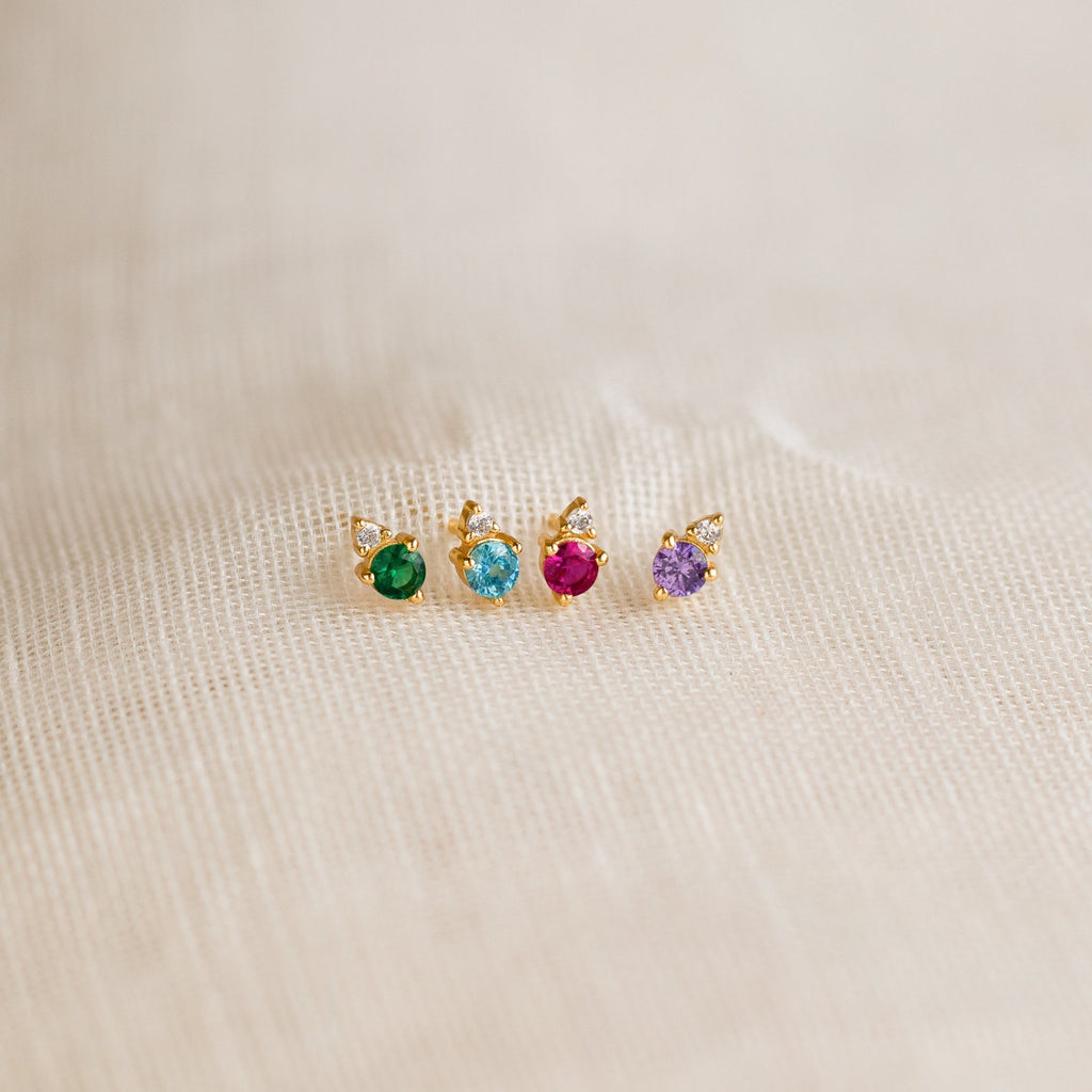 Four dainty Raindrop Birthstone Studs featuring green, blue, pink, and purple stones shine on beige fabric—perfect colorful earrings to add a touch of elegance to your jewelry collection.