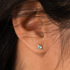 Close-up of an ear adorned with Raindrop Birthstone Studs—dainty earrings featuring a small blue gemstone stud with a tiny clear stone above, perfect for lovers of subtle birthstone jewelry.