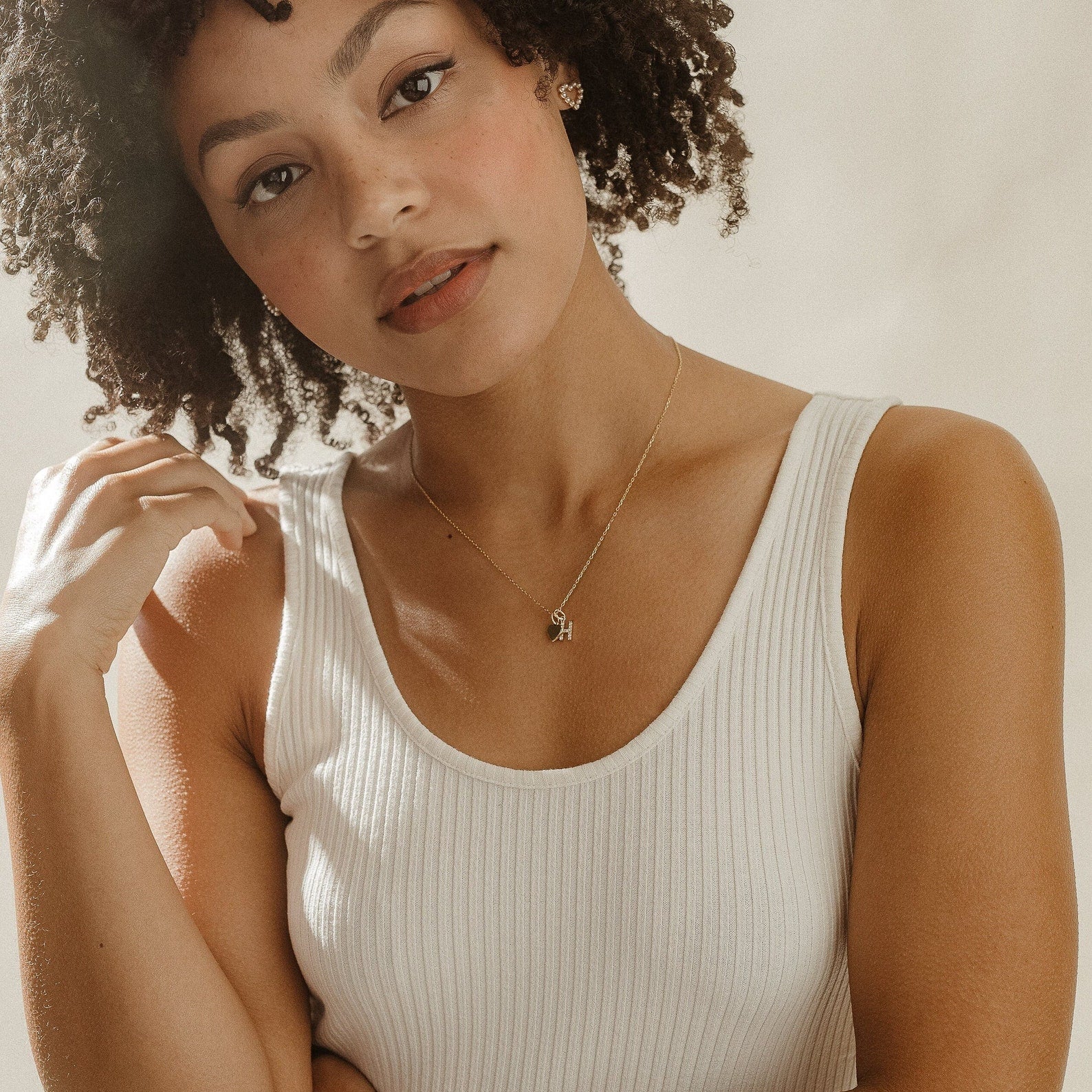 A woman with curly hair wears a white tank top and the Pave Initial Heart Necklace, gazing softly at the camera.