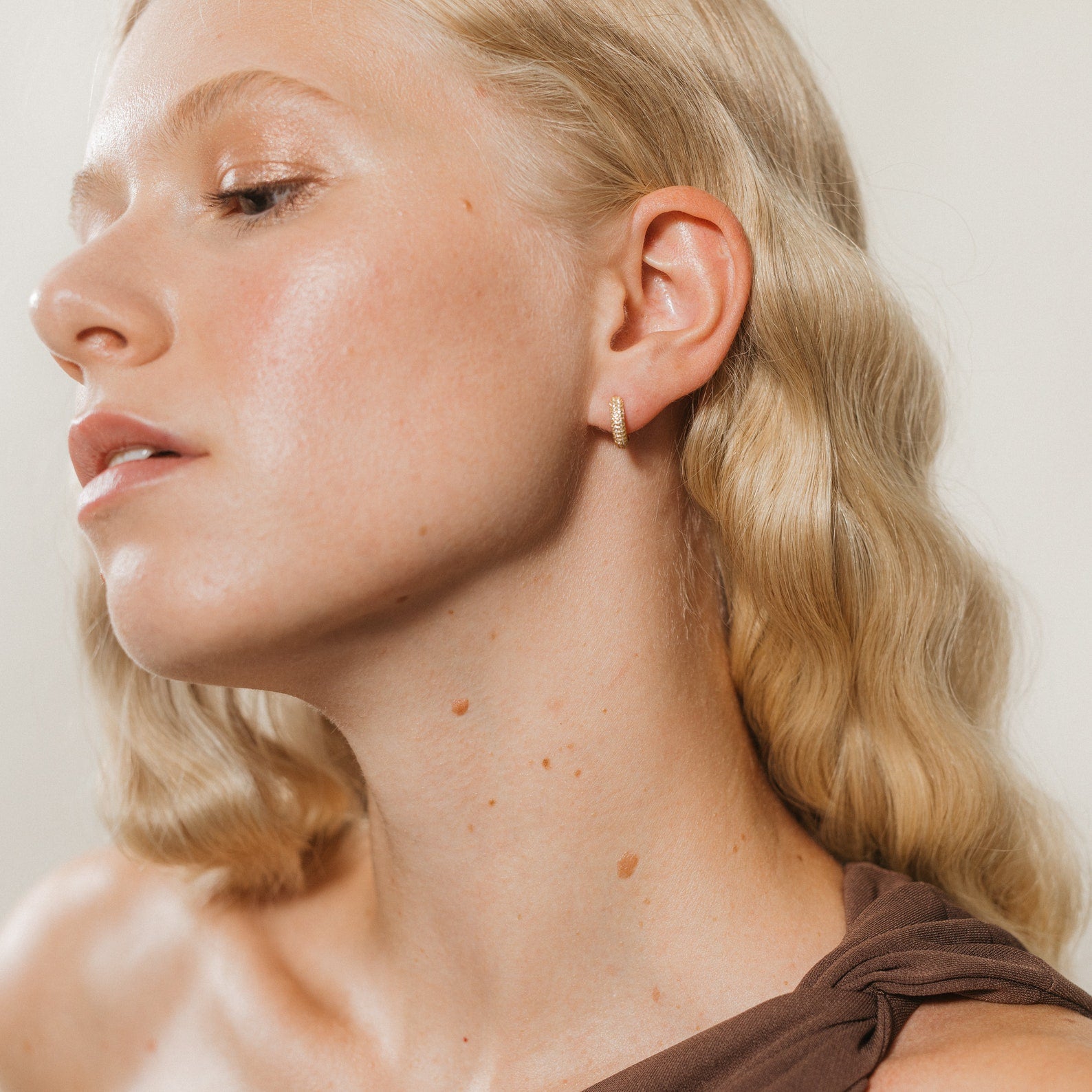 Woman with wavy blonde hair, dressed in a brown top and wearing Clarissa Pave Huggies, looks to the side.