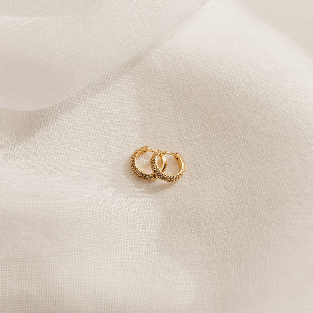 The Clarissa Pave Huggies—two small gold earrings with textured detailing—rest on soft, white fabric, perfect for adding a subtle sparkle to your date night jewelry collection.