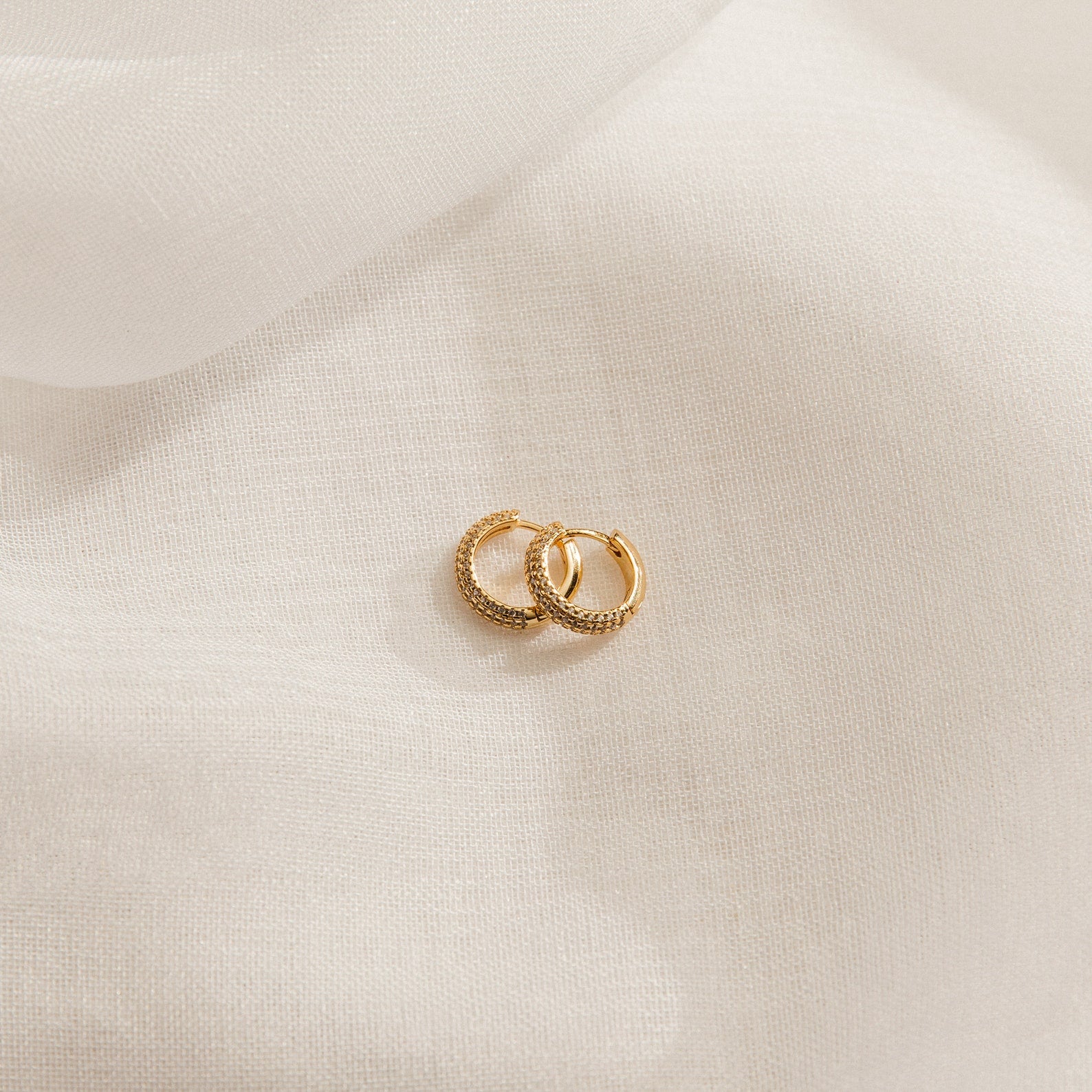 The Clarissa Pave Huggies—two small gold earrings with textured detailing—rest on soft, white fabric, perfect for adding a subtle sparkle to your date night jewelry collection.