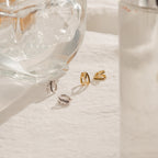 The Amelia Onyx Duo Huggies—two gold and two silver minimalist earrings—are displayed on a white cloth next to a clear glass ornament and a glass of water.