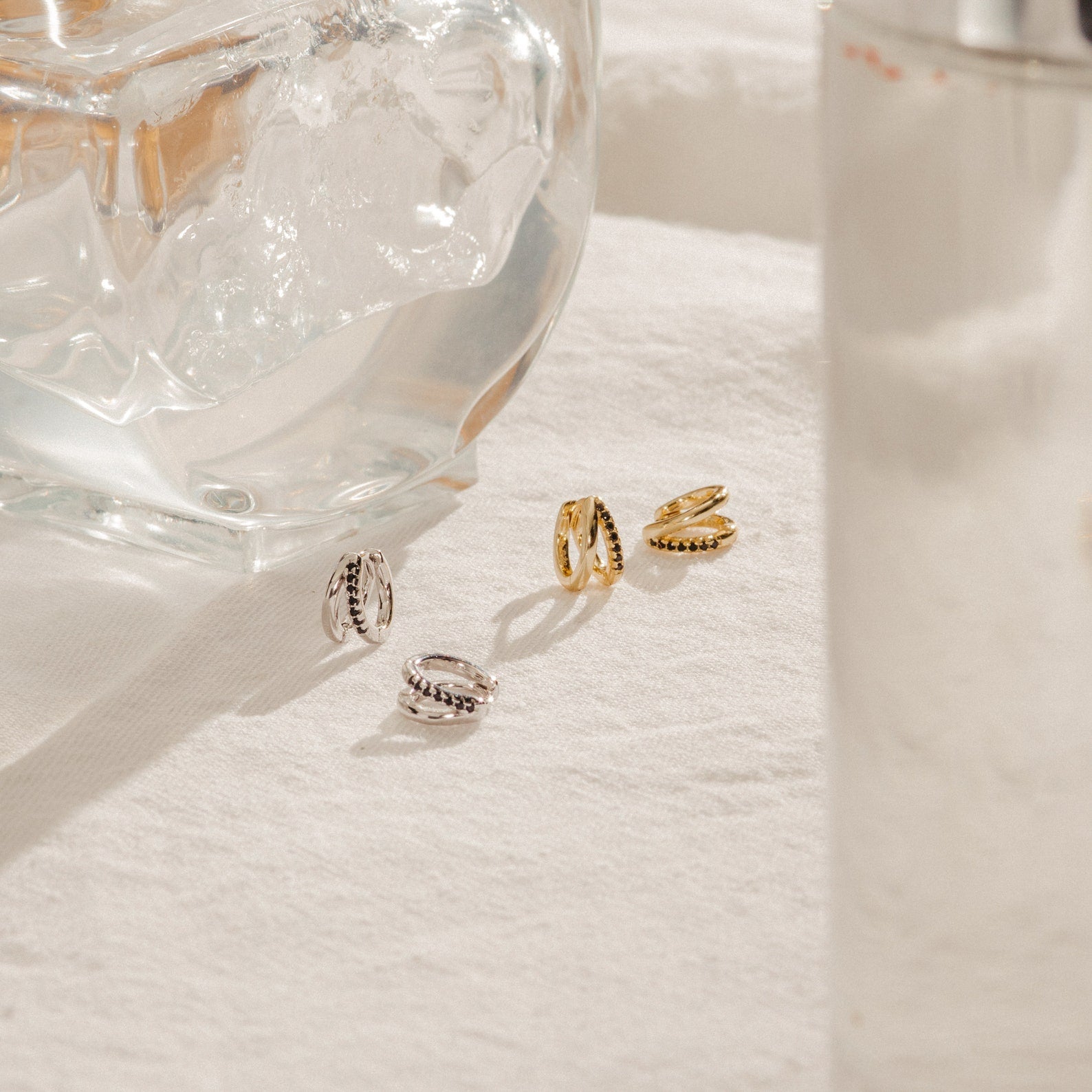 The Amelia Onyx Duo Huggies—two gold and two silver minimalist earrings—are displayed on a white cloth next to a clear glass ornament and a glass of water.
