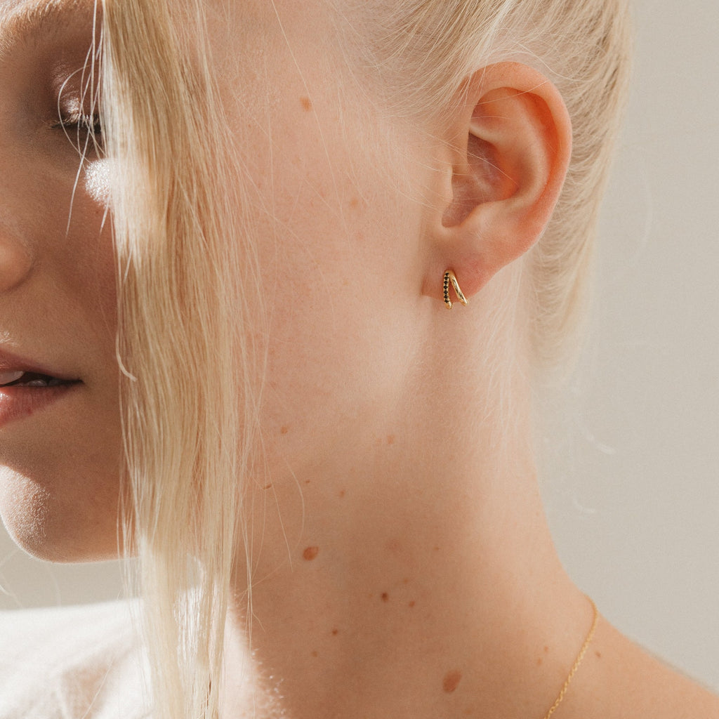 Close-up of a woman’s ear with Amelia Onyx Duo Huggies in gold, blonde hair, light skin, and a thin gold necklace.