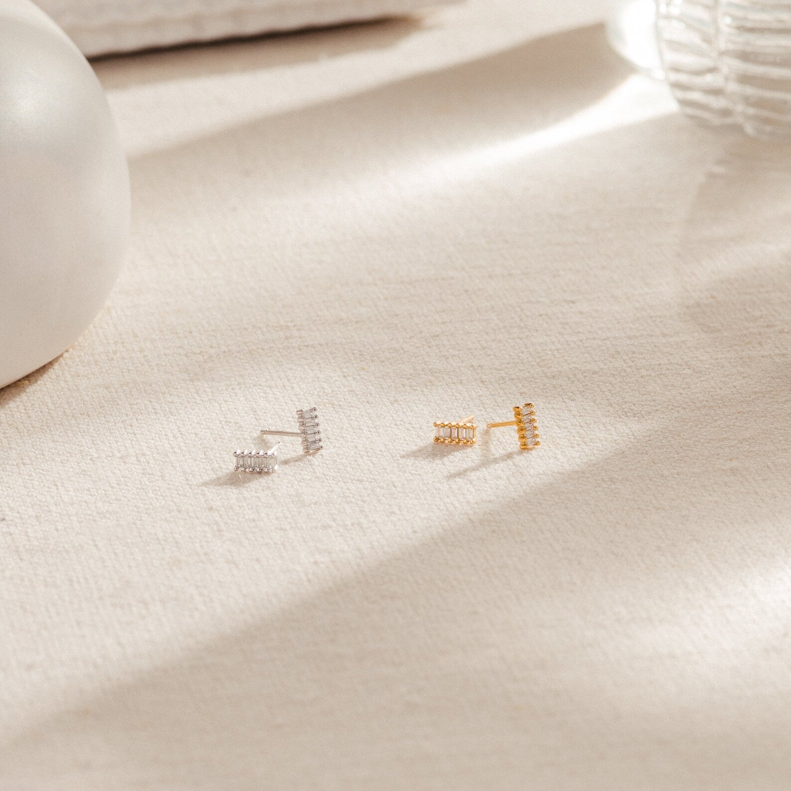Two pairs of Serena Baguette Studs in silver and gold, displayed on a light textured fabric surface.