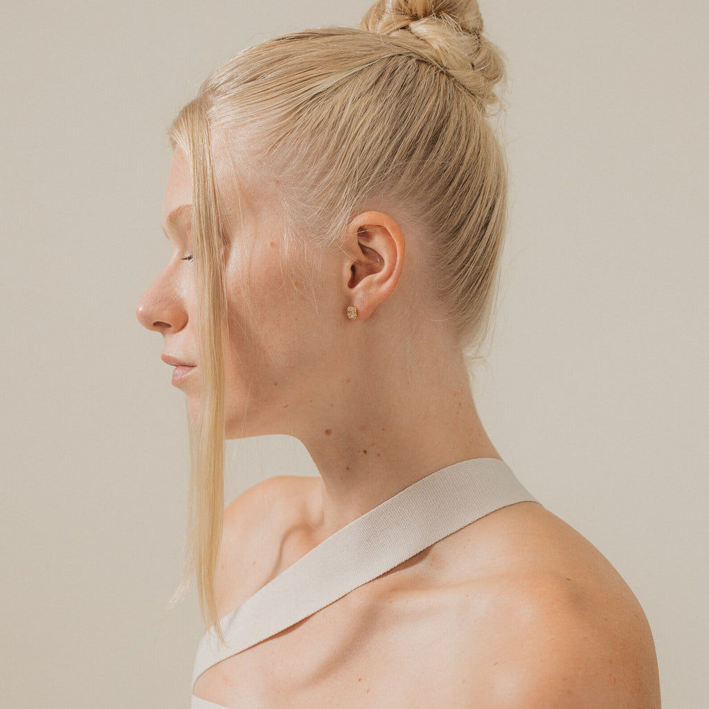 A woman with blonde hair in a bun, wearing a white top and the elegant Arabella Diamond Studs, is seen in profile against a plain background.