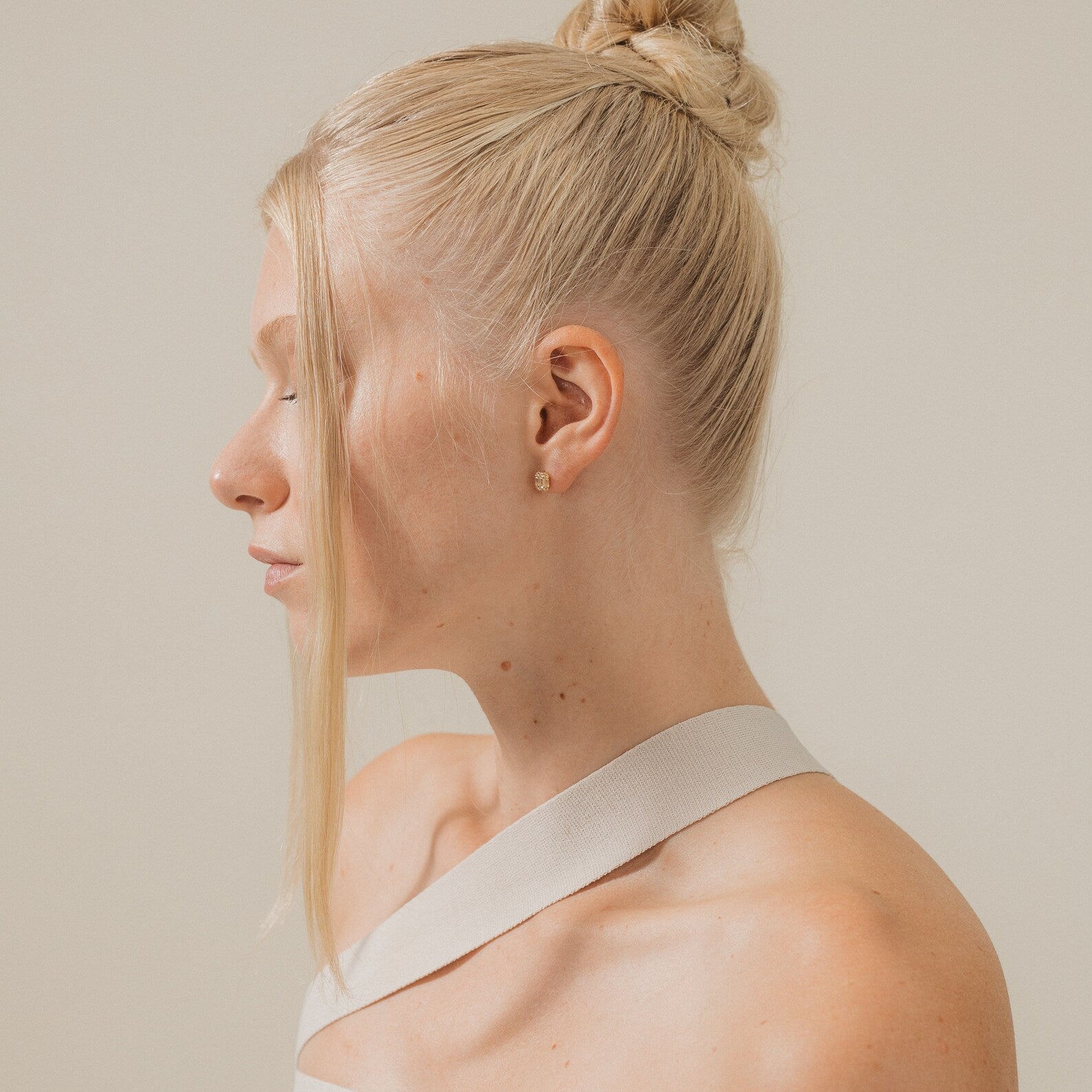 A woman with blonde hair in a bun, wearing a white top and the elegant Arabella Diamond Studs, is seen in profile against a plain background.