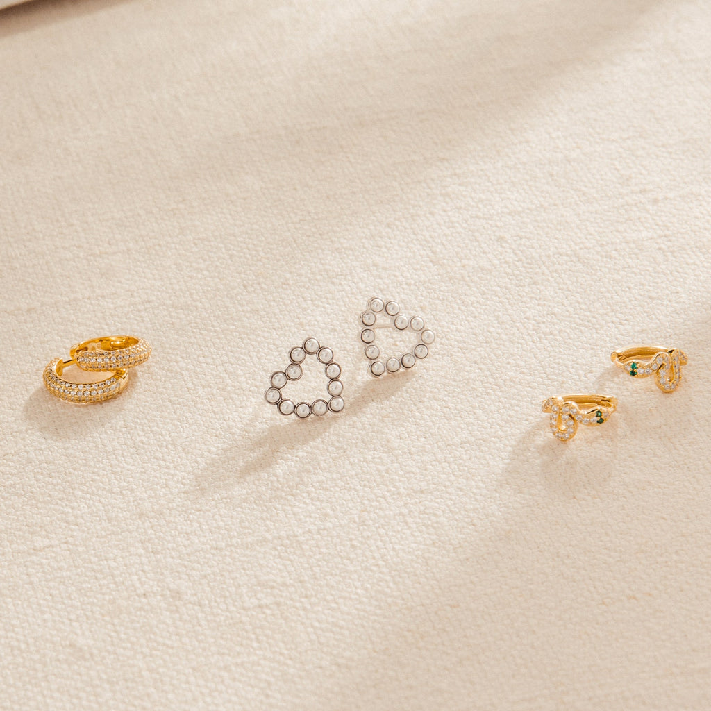 Three pairs of earrings—gold hoops, silver heart shapes, gold studs—and a pair of Pearl Heart Studs with dainty beaded charms are displayed on light fabric, adding a touch of vintage jewelry elegance.