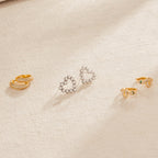 Three pairs of earrings—gold hoops, silver heart shapes, gold studs—and a pair of Pearl Heart Studs with dainty beaded charms are displayed on light fabric, adding a touch of vintage jewelry elegance.