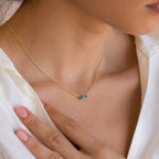 A woman wearing a single Emery Birthstone Necklace in gold with a bright Blue Zircon rectangular gemstone, styled against a white blouse.