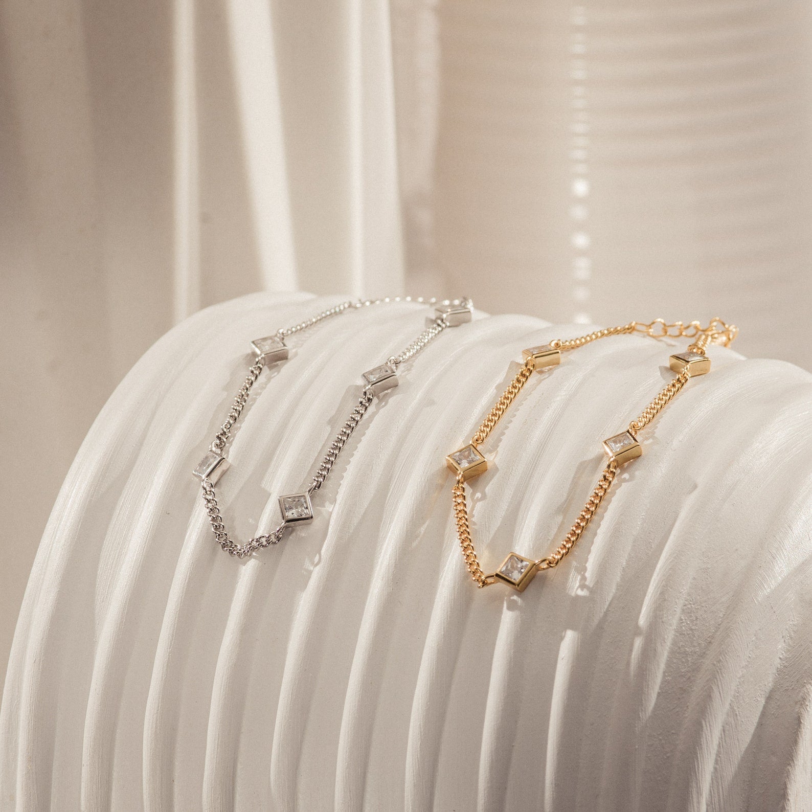 The Venus Diamond Station Bracelets—one in silver, one in gold—each feature sparkling square crystal charms, making them perfect as wedding or anniversary jewelry, elegantly showcased on a white surface.