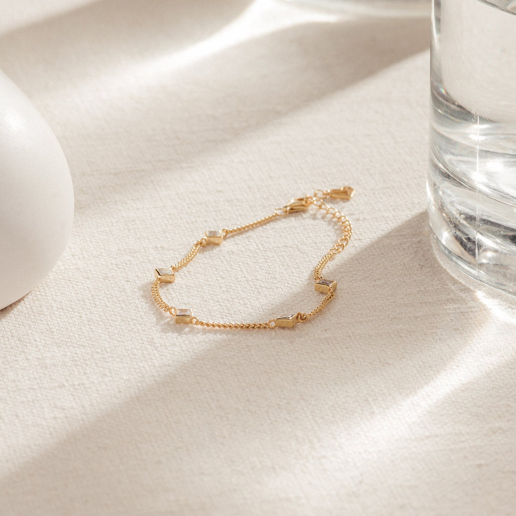 The Venus Diamond Station Bracelet—a delicate gold chain with diamond accents—rests on cream fabric beside a glass of water. An elegant choice for wedding jewelry or an unforgettable anniversary gift.