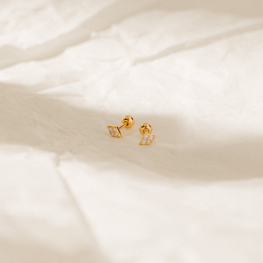 The Jocelyn Diamond Studs are dainty earrings with square, gem-encrusted gold studs, shown on a crumpled beige background.